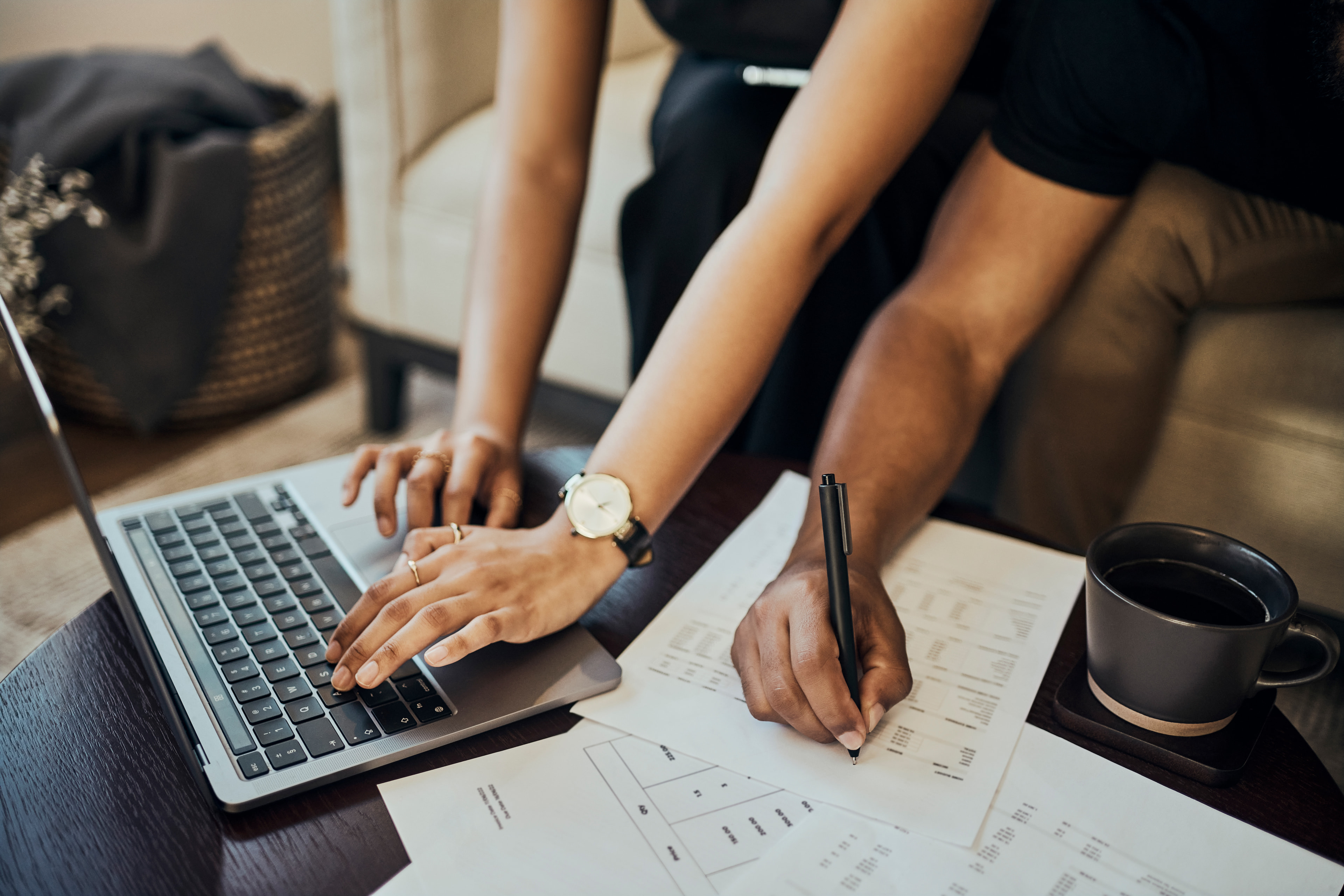 A close-up, high-angle shot of two people collaborating at a dark wooden coffee table. One person’s hands are typing on a silver laptop, while the other person uses a black pen to write on financial documents. A grey coffee mug on a coaster and several scattered papers with charts and tables are also visible.